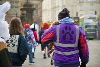 People, a mix of fursuiters and none, walking away from the camera. In the foreground is a person wearing a purple, BristolFurs-branded hi-vis vest.