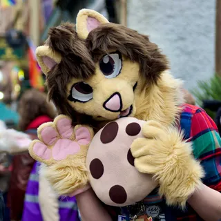 A fursuiter of a brown cat holding a large, plushie cookie to its mouth and waving at the viewer.